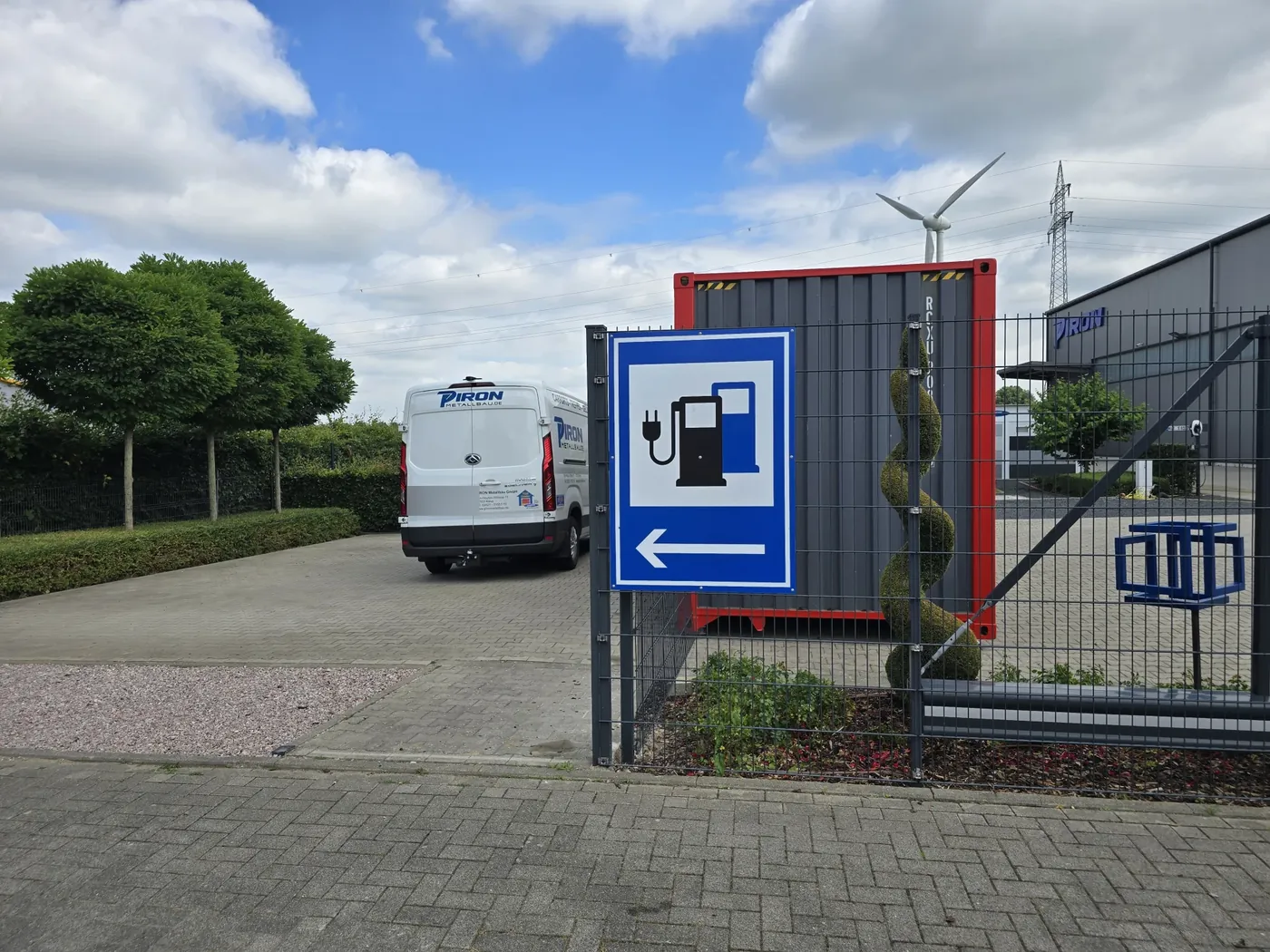 Blaues Ladesäulen-Hinweisschild mit Pfeil nach links am Zaun der Zufahrt, dahinter Container, Firmenwagen und die PIRON-Halle
